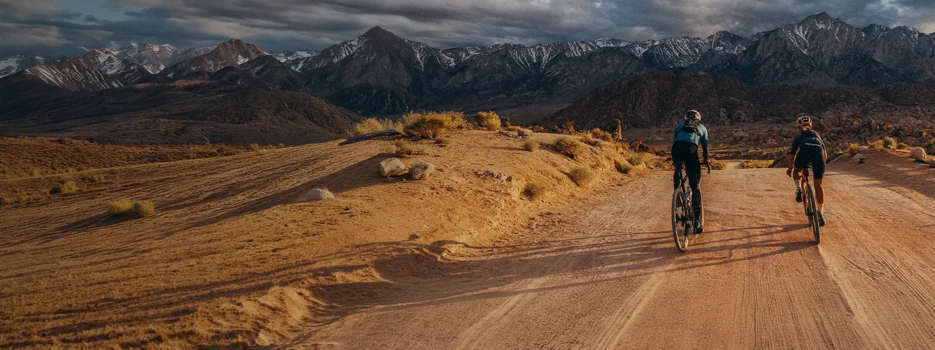 Two cyclists riding along an open gravel road towards a mountain range