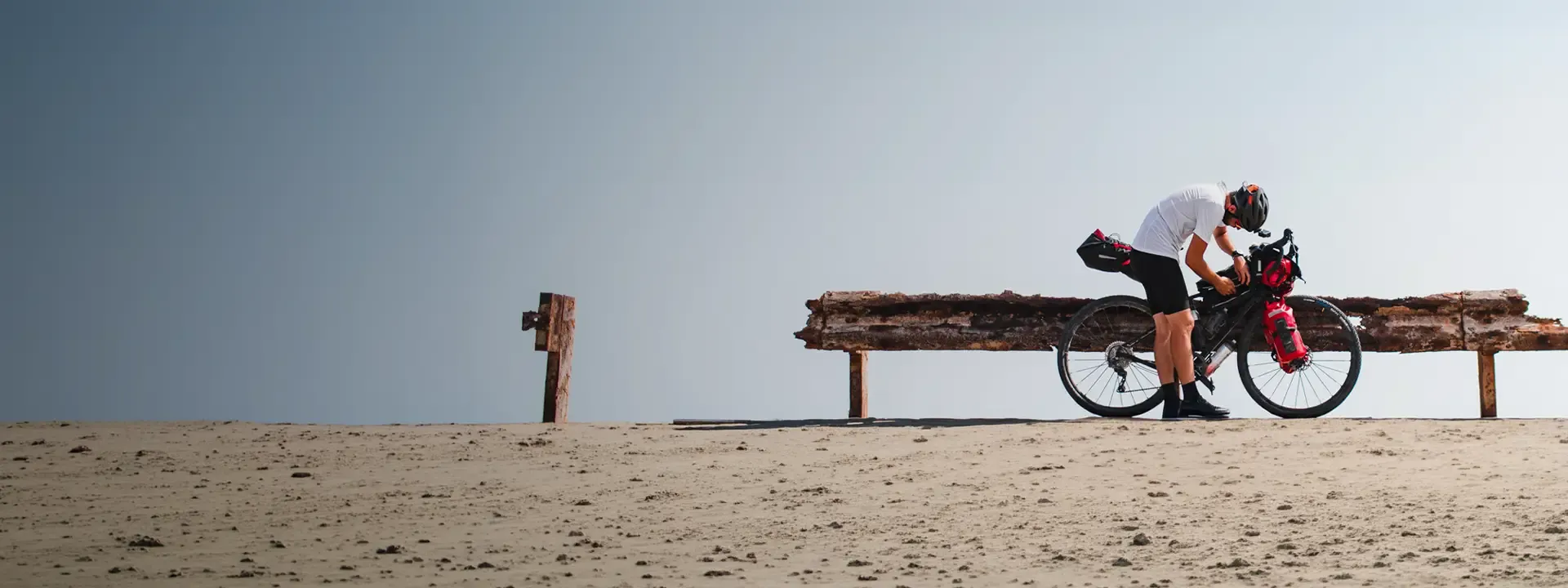 Cyclist checking a luggage-equipped bike beside a wooden barrier on a sandy track
