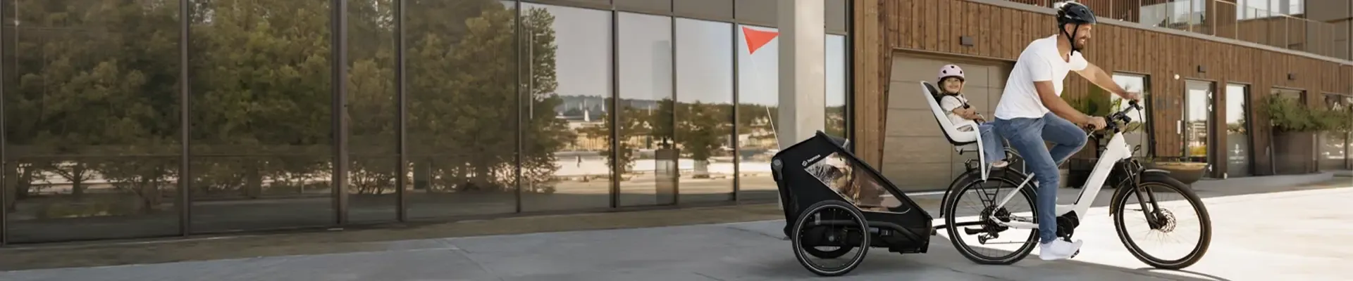 Father riding an electric bike with a child in a rear child seat and another child in a bike trailer.