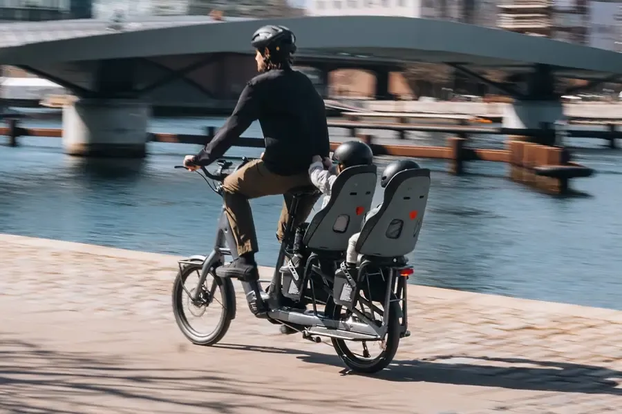 Parent riding a cargo e-bike with two helmeted children in rear-mounted child seats.