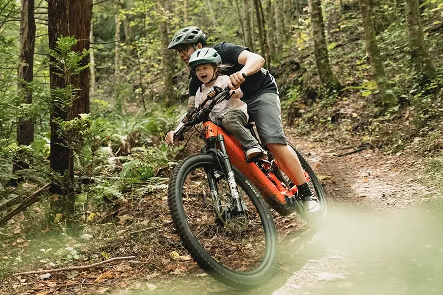 Parent and helmeted child riding a mountain bike with a top-tube-mounted child seat on a forest trail.