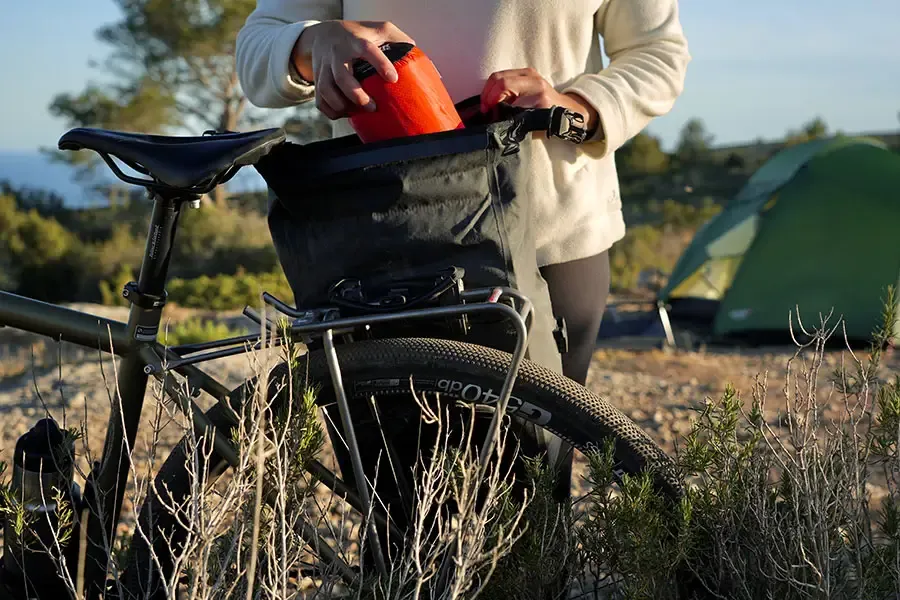 Cyclist packing equipment into a pannier bag mounted on a rear bike rack at a campsite.