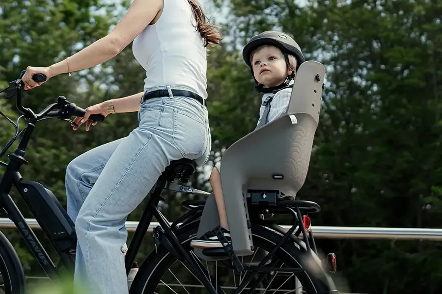 Parent cycling with a helmeted child in a rack-mounted rear bike seat.
