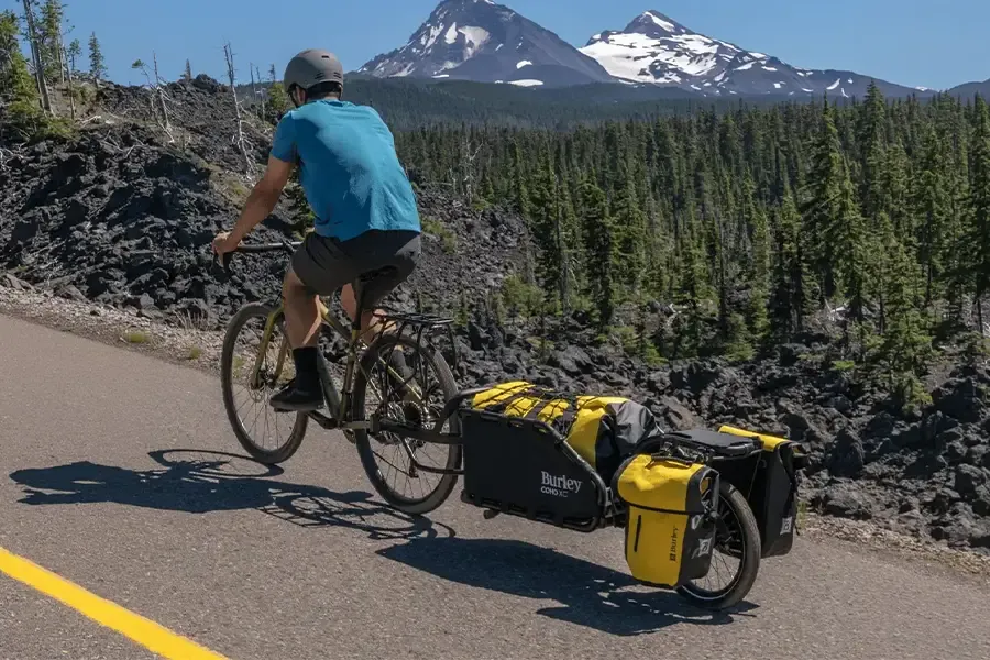 Cyclist towing a loaded cargo trailer with pannier bags on a mountain road.