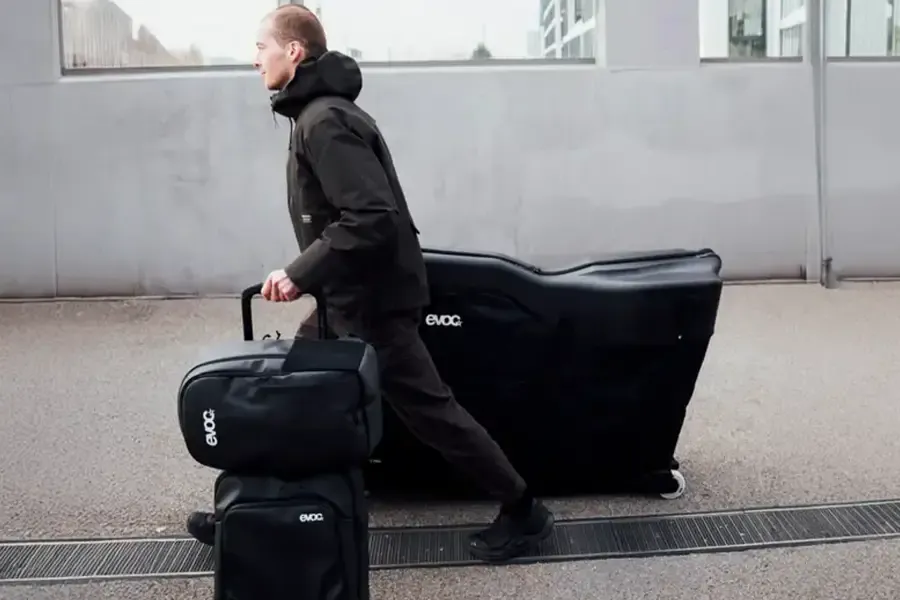 man walking with a bike in an EVOC Road Bike Bag Pro and wheeled luggage